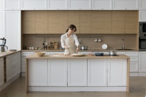 A woman assembles a pizza on her kitchen island. You can see a variety of toppings and pizza dough scattered across the table.