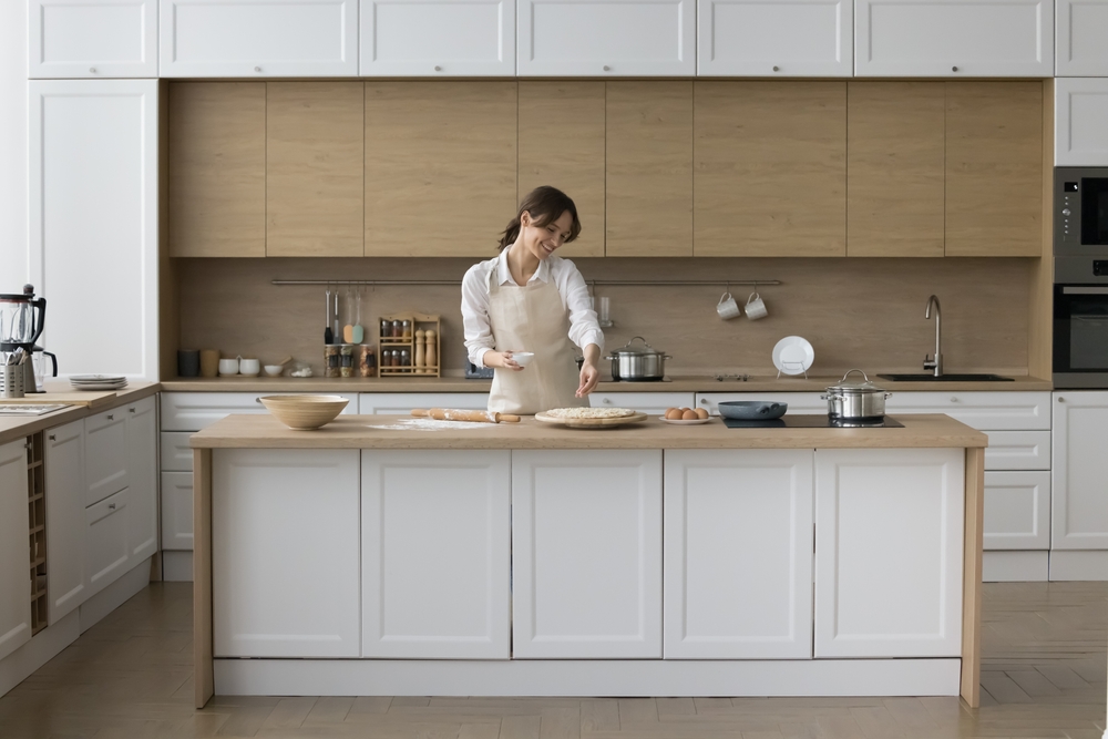 A woman assembles a pizza on her kitchen island. You can see a variety of toppings and pizza dough scattered across the table.
